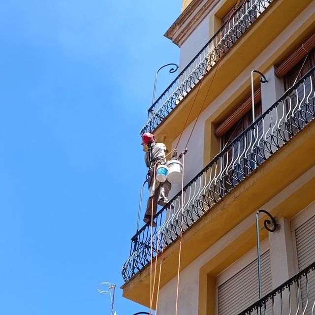 Hombre pintando el exterior de un edificio suspendido por cuerdas, día soleado.