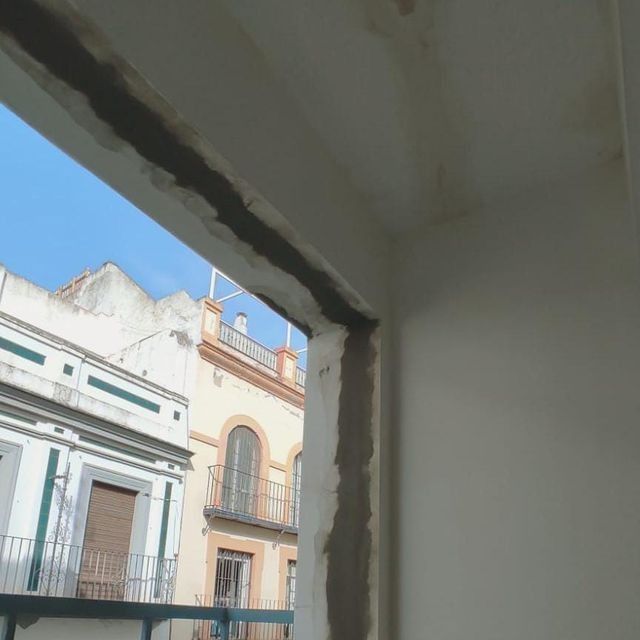 Vista desde una ventana que da a edificios con balcones y cielo azul. El marco de la ventana tiene cemento a la vista.