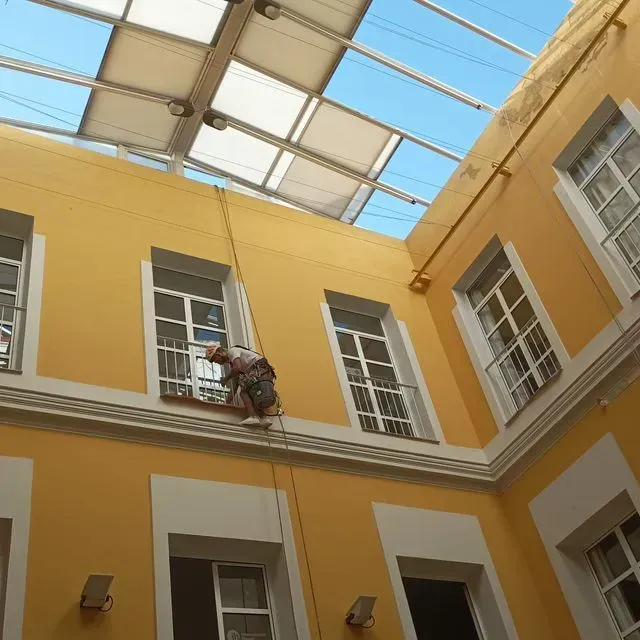 Un trabajador limpia una ventana en la fachada amarilla de un edificio, sostenido por cuerdas. Se ve el tragaluz y el cielo.