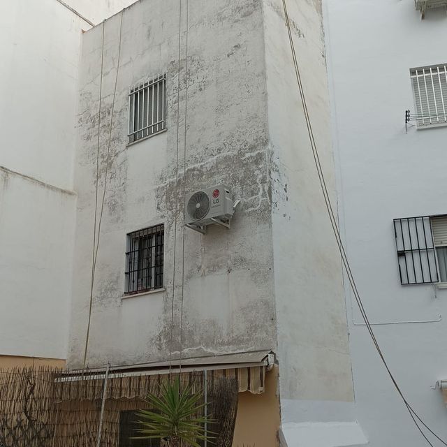 Edificio estrecho de color blanco con dos ventanas enrejadas y aire acondicionado, situado entre otros dos edificios.