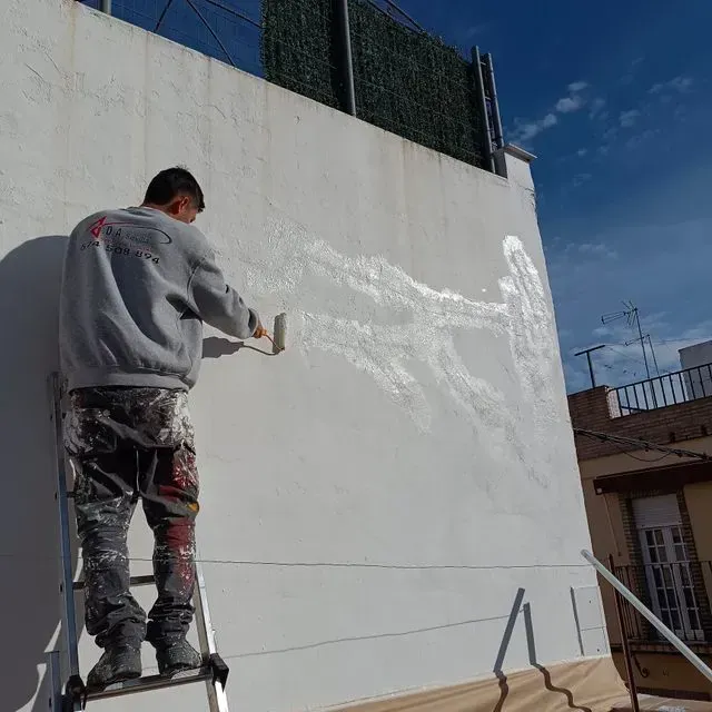 Persona subida a una escalera pintando una pared blanca con un rodillo. Al aire libre, con luz natural brillante.