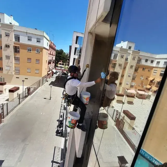 Un limpiador de ventanas desciende en rappel por un edificio, limpiando una gran ventana con edificios de la ciudad al fondo.