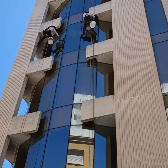 Limpiadores de ventanas limpian la fachada de cristal de un edificio alto, suspendidos por cuerdas. El edificio es de hormigón y de líneas angulares.