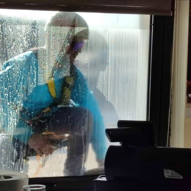 Persona con chaqueta azul limpiando una ventana con una rasqueta. La ventana está mojada y tiene una persiana enrollable encima.