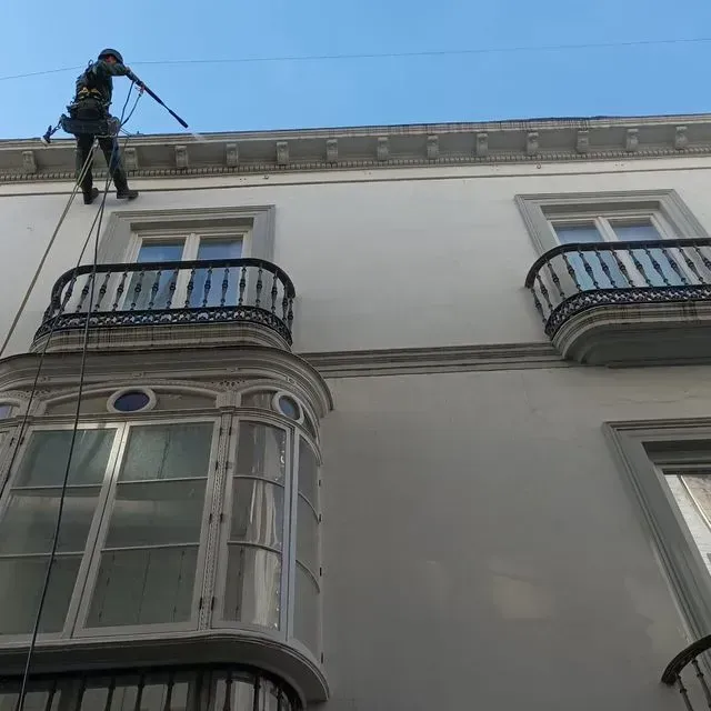 Una persona sujeta con cuerdas realiza una limpieza a presión de un edificio blanco con balcones.