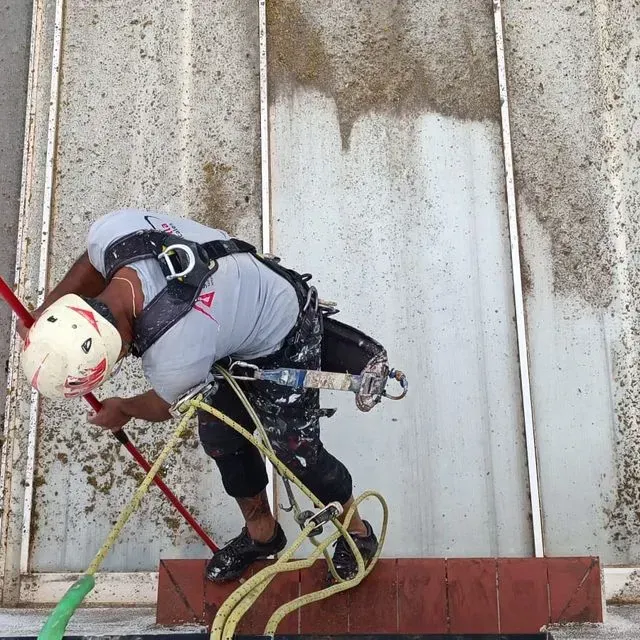 Hombre con equipo de seguridad, limpiando una pared desde altura con una manguera.