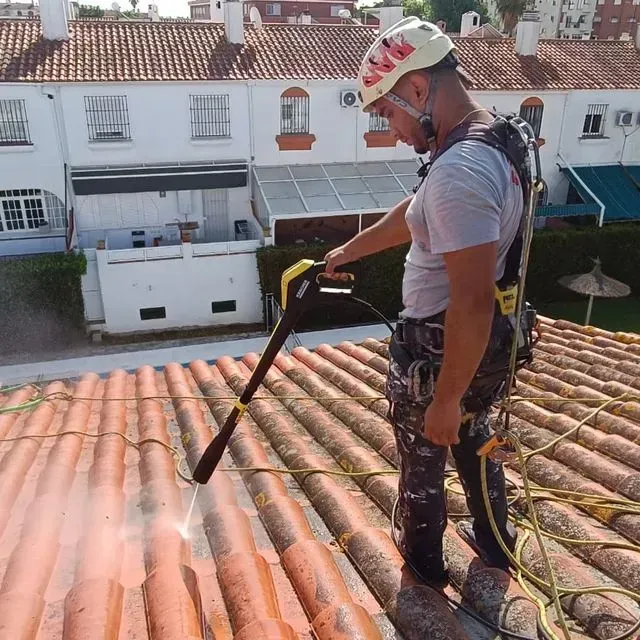 Hombre lavando a presión un tejado de tejas de terracota, sujeto con arnés de seguridad.