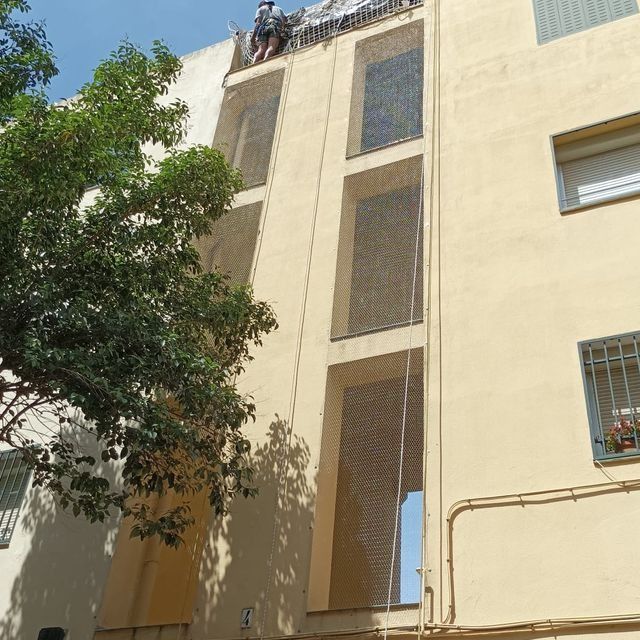 Unos obreros subidos a un andamio instalan redes en la fachada de un edificio alto para proteger las zonas con ventanas abiertas.