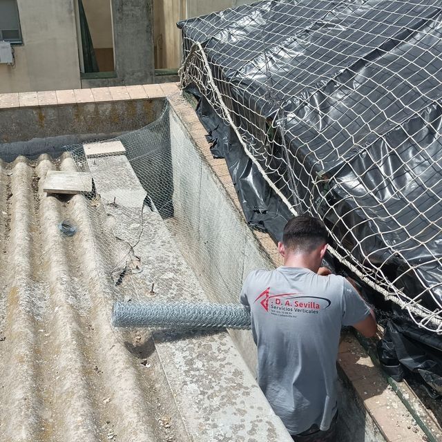 Un hombre instala una malla metálica en un tejado cerca de una zona cubierta con una lona negra.