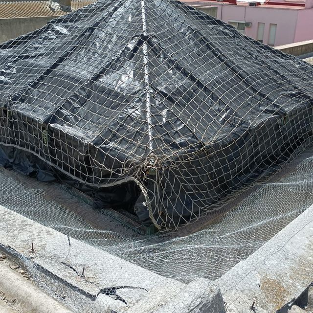 Lona y red negras cubren una estructura cuadrada en una azotea. Edificio gris al fondo.