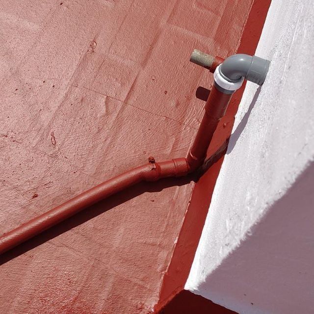 Techo pintado de rojo con una tubería de drenaje roja conectada a una tubería gris y pared blanca.