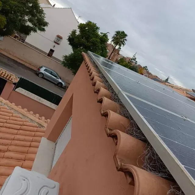 Paneles solares sobre un tejado de tejas de terracota, con una calle y edificios al fondo. Cielo nublado.