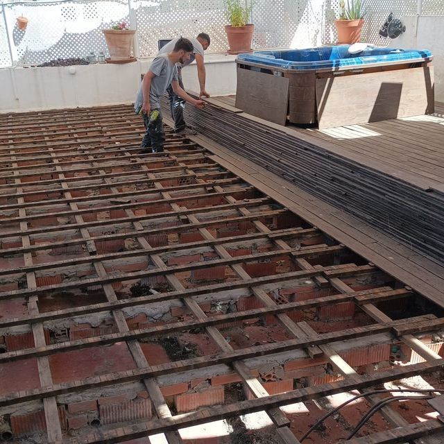 Dos personas instalando tablas de terraza en un patio de azotea con jacuzzi. Se ven la estructura de madera y los ladrillos a la vista.