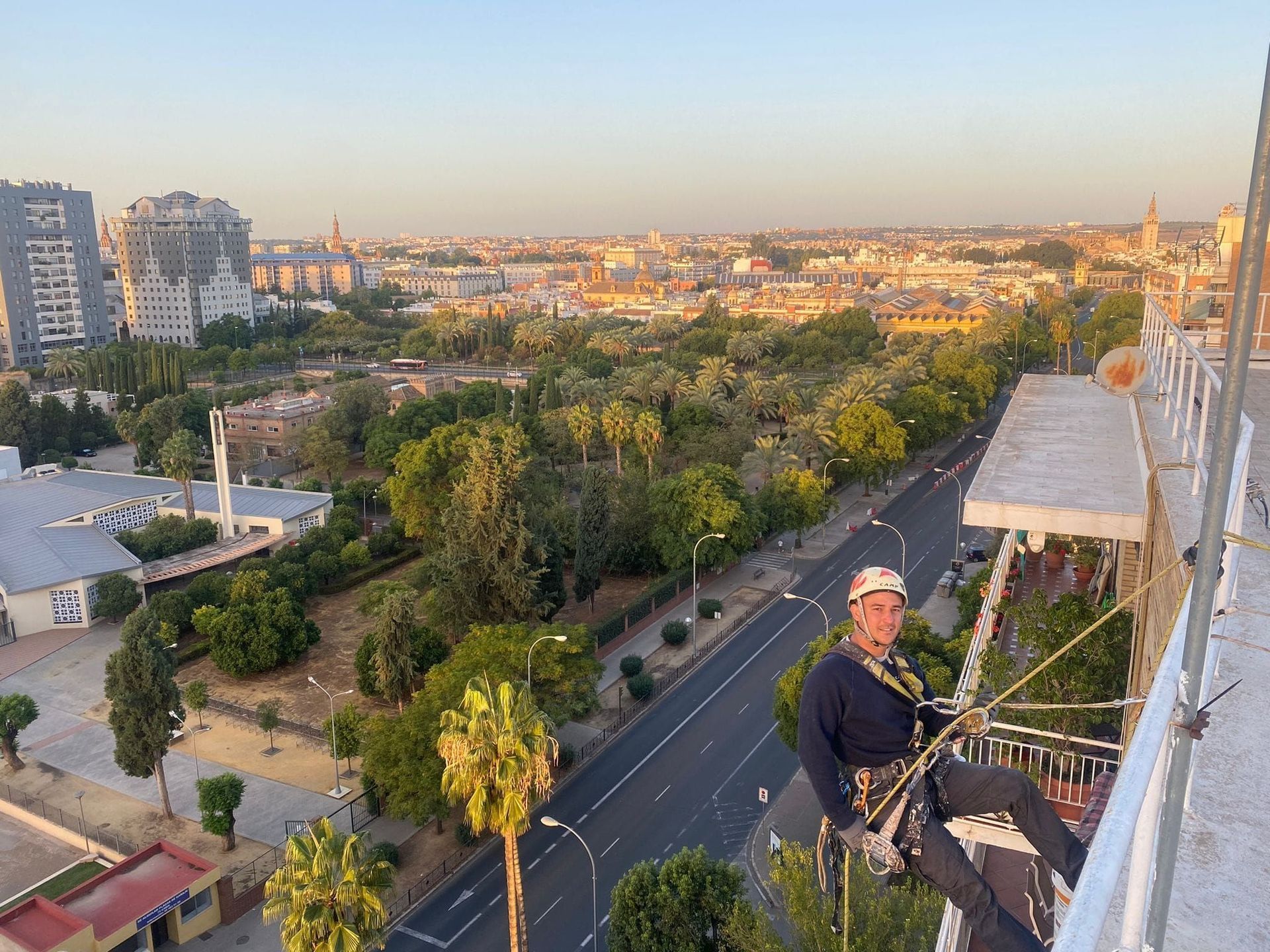 Persona descendiendo en rappel por un edificio, con vistas a una ciudad con árboles verdes y una carretera.