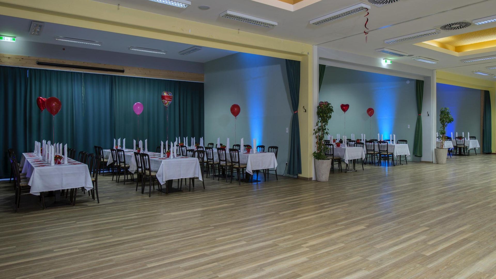 Empty banquet hall with round tables, white tablecloths, red balloons, and blue curtains along the wall