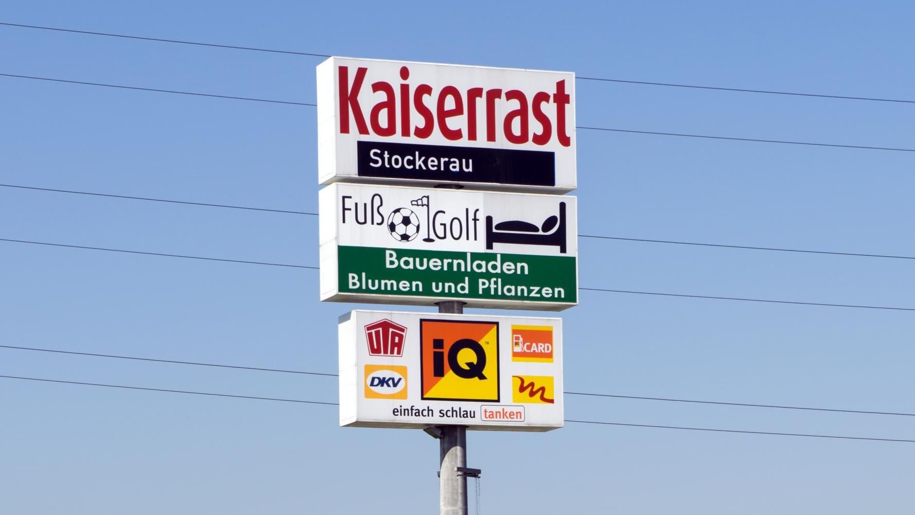 Roadside sign for Kaiserast Stuckerei and garden supplies against a blue sky.