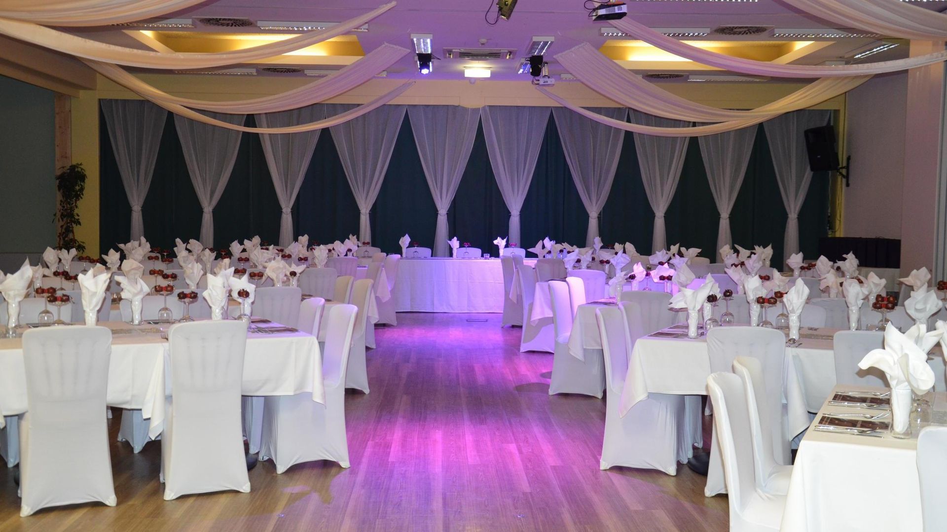 Elegant banquet hall with round white-clothed tables, chairs, and purple accent lighting at the center aisle
