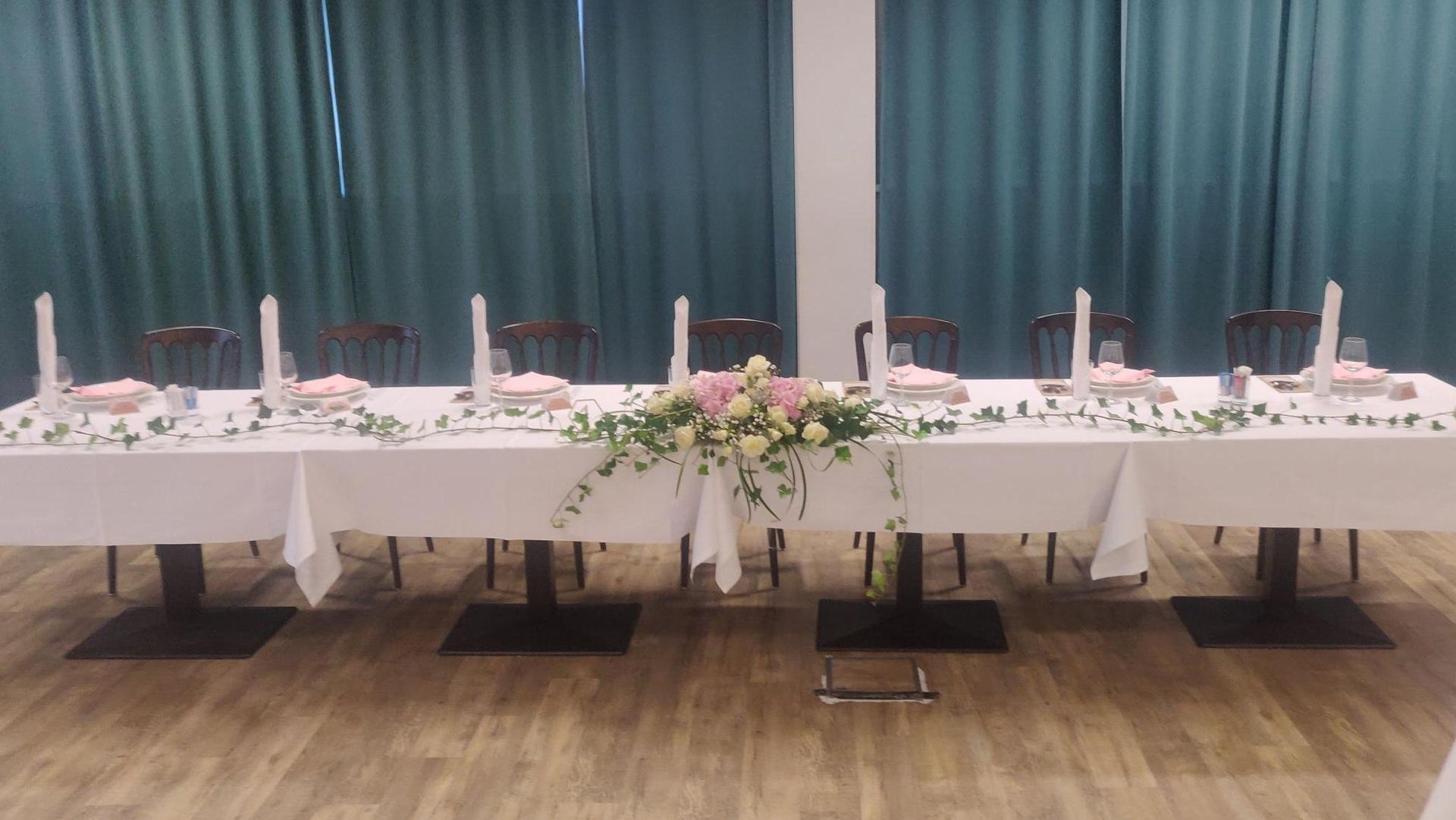 Long banquet table with pink floral centerpieces, white tablecloth, and teal curtains in a decorated hall
