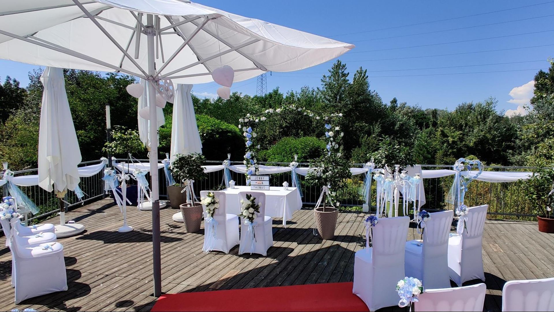 Outdoor patio with white umbrellas and table settings, decorated for an event under a sunny blue sky