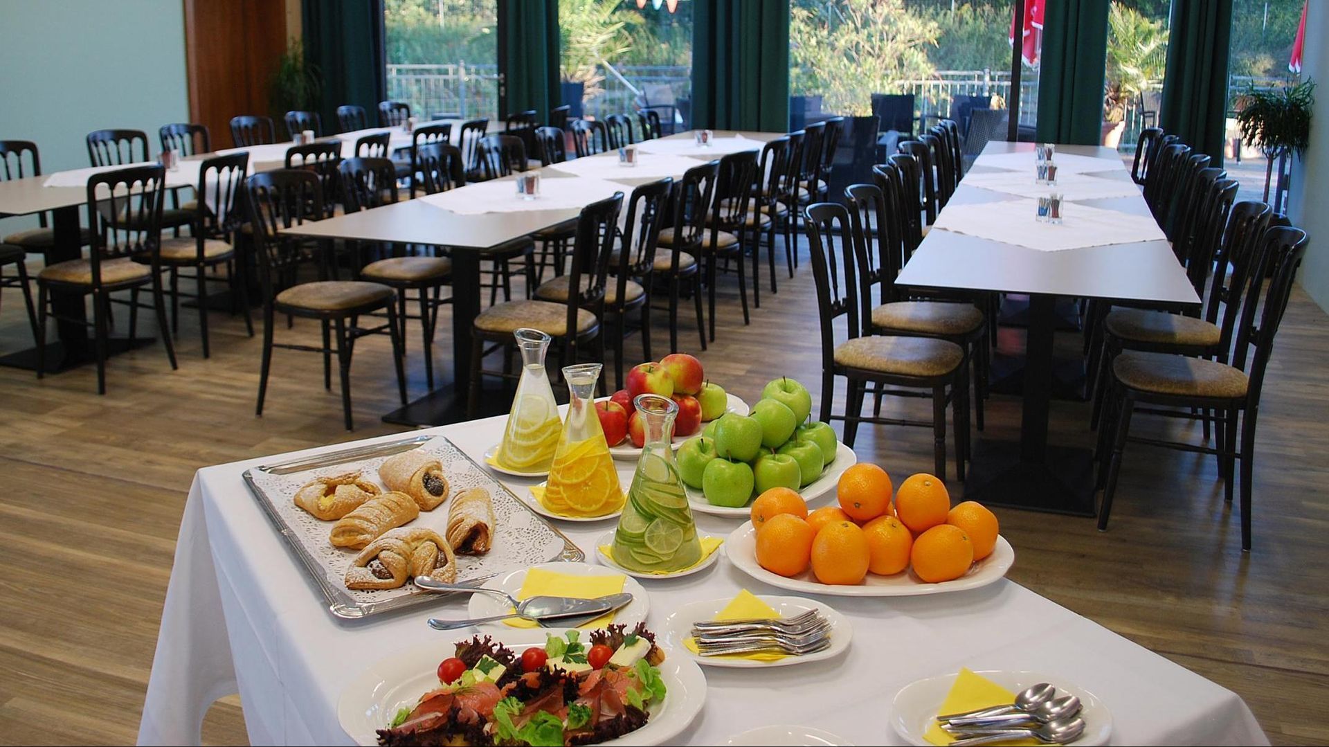 Buffet table with fruit, desserts, and appetizers in a bright dining hall with rows of tables and chairs