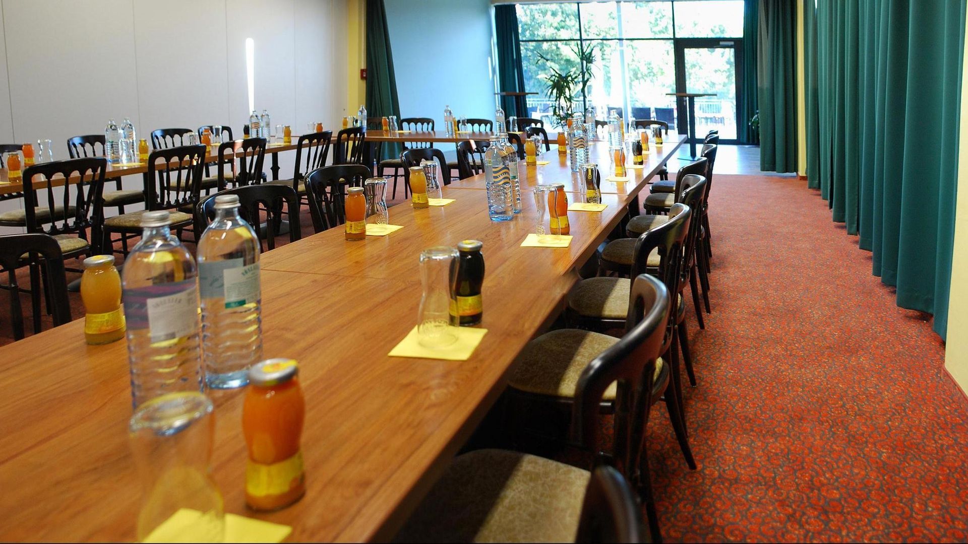 Long wooden table set with bottles and glasses in a bright dining hall with chairs and green curtains.