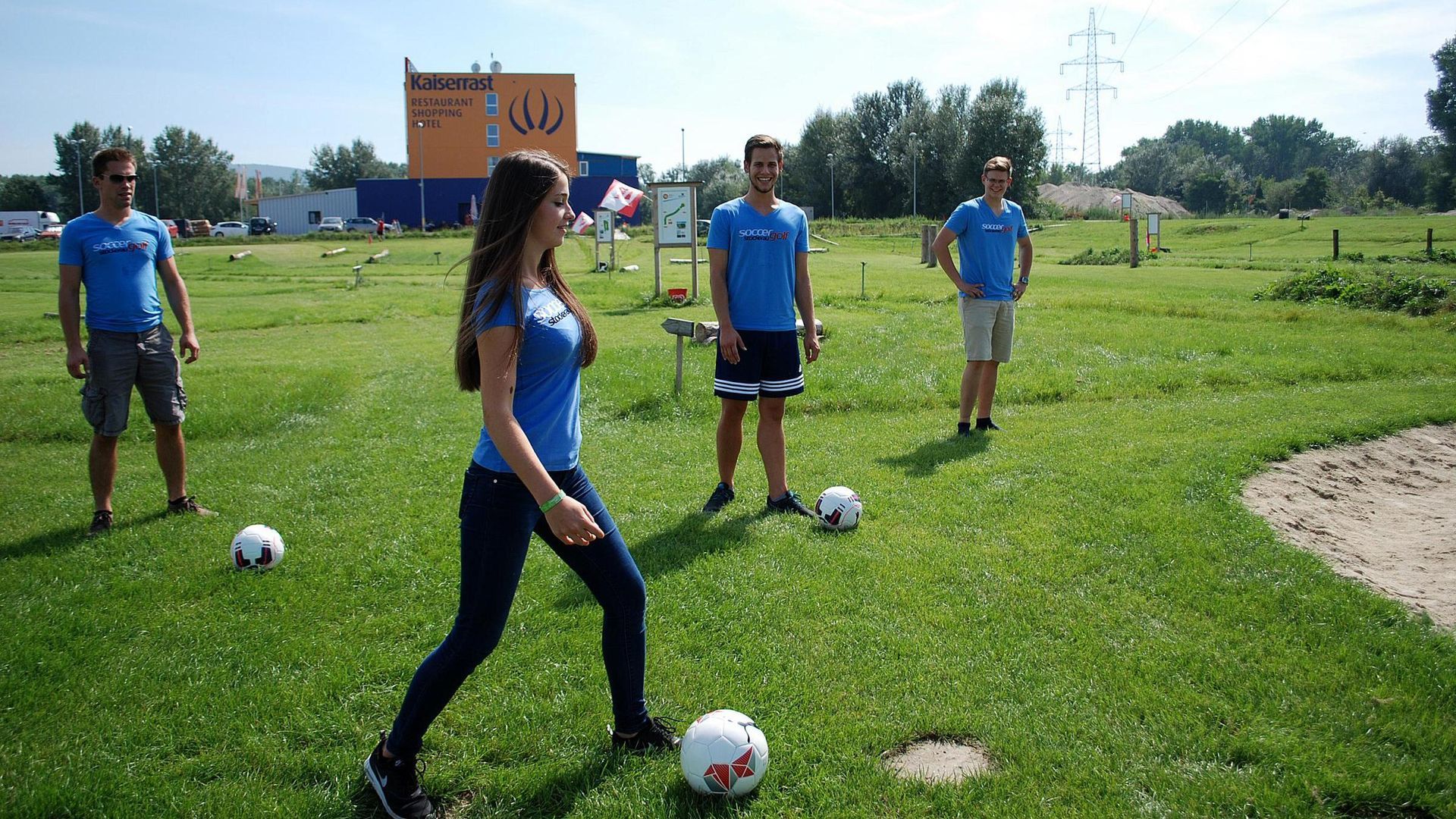 People in blue sportswear standing on a grassy soccer field with balls during practice