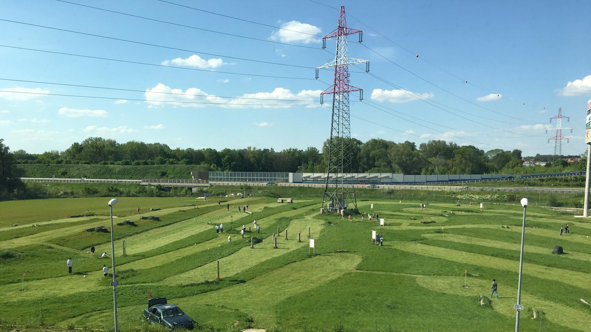 Green park landscape with people walking, power lines overhead, and a red transmission tower under a blue sky