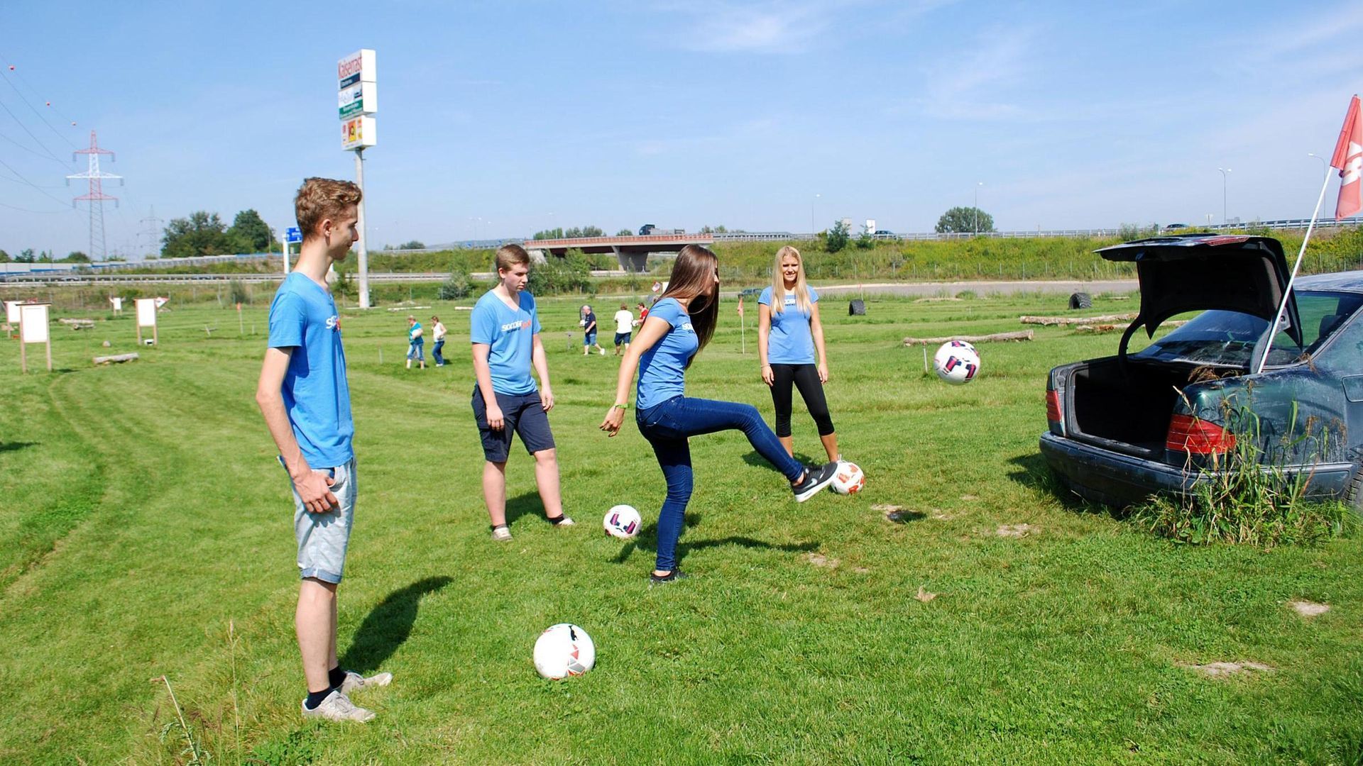 People playing soccer on a grassy field near an open car trunk with sports gear.