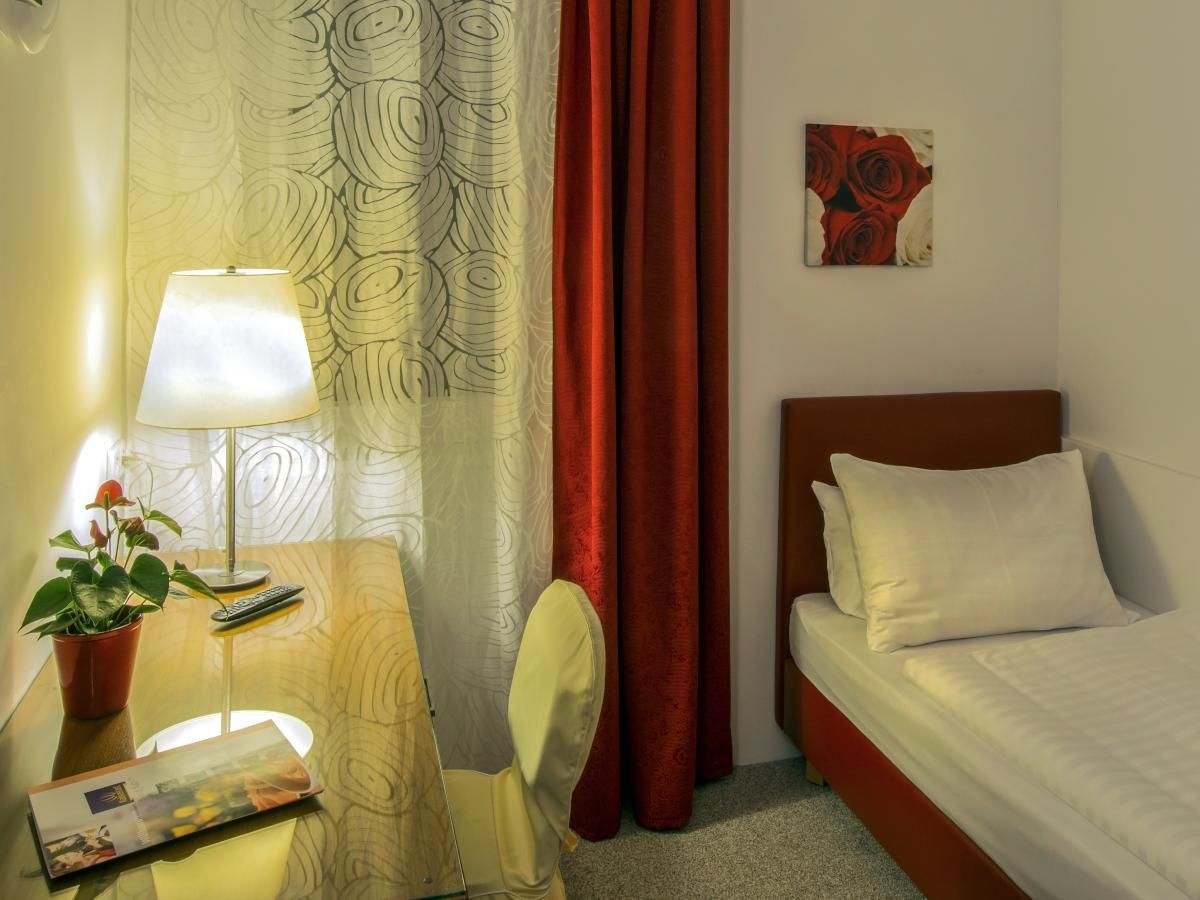 Small cozy bedroom with a single bed, desk lamp, chair, curtains, and a small wall painting.
