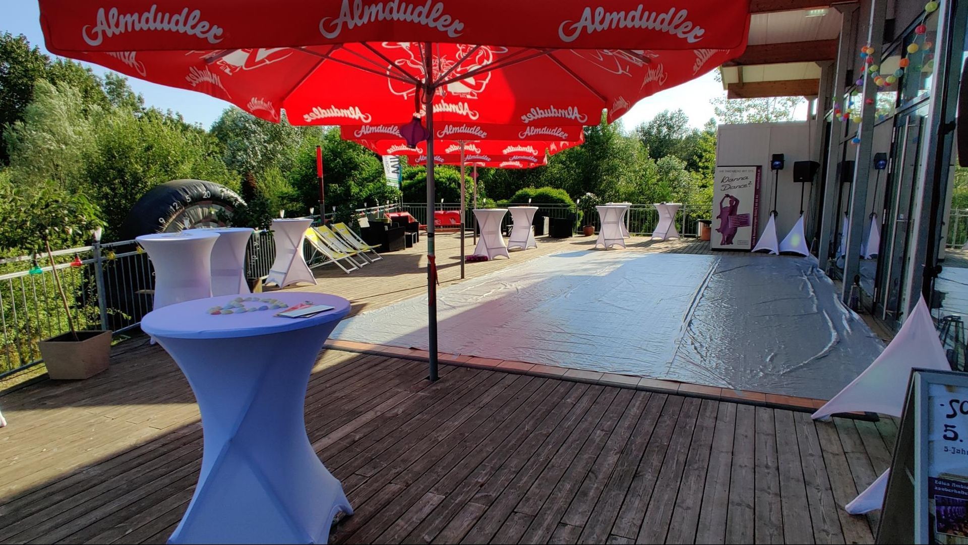 Outdoor event setup with red umbrellas, high table, wooden deck, and white gravel path by storefronts.