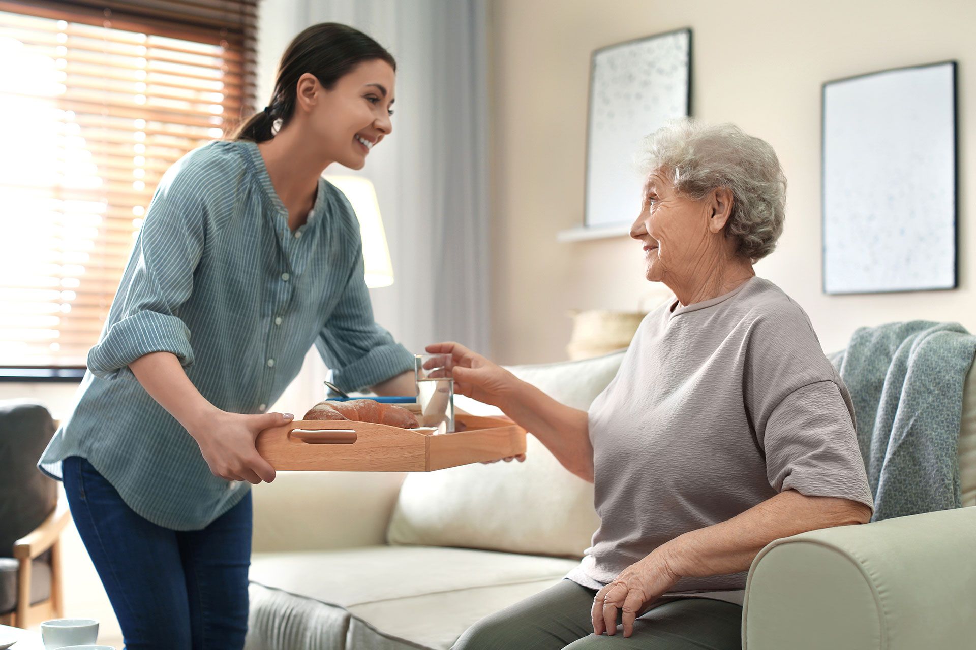 In einem Wohnzimmer gibt eine Frau einer älteren Frau ein Tablett mit Essen.