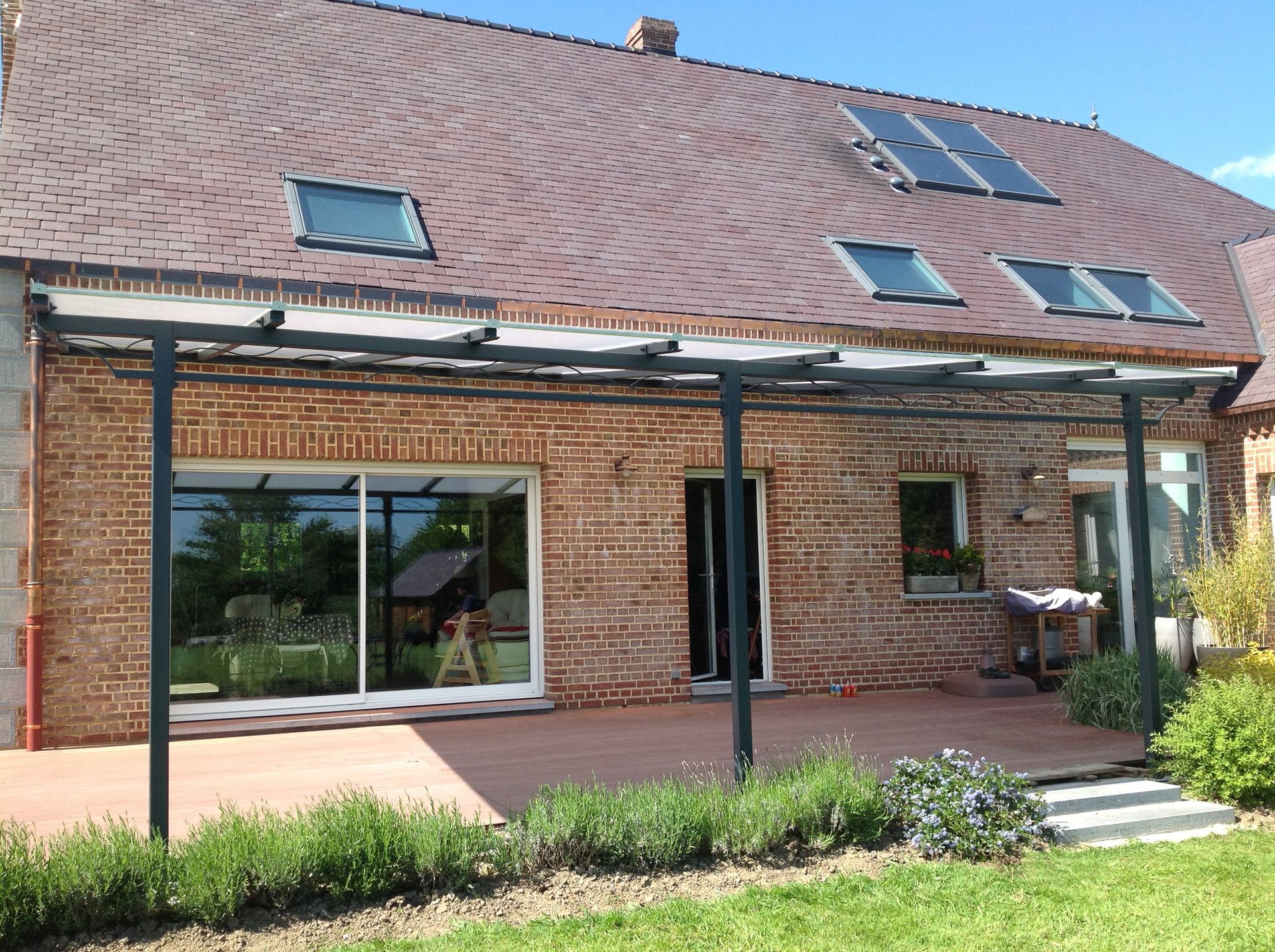 Patio moderne avec pergola en métal noir, terrasse en bois et verdure à côté d'une maison blanche