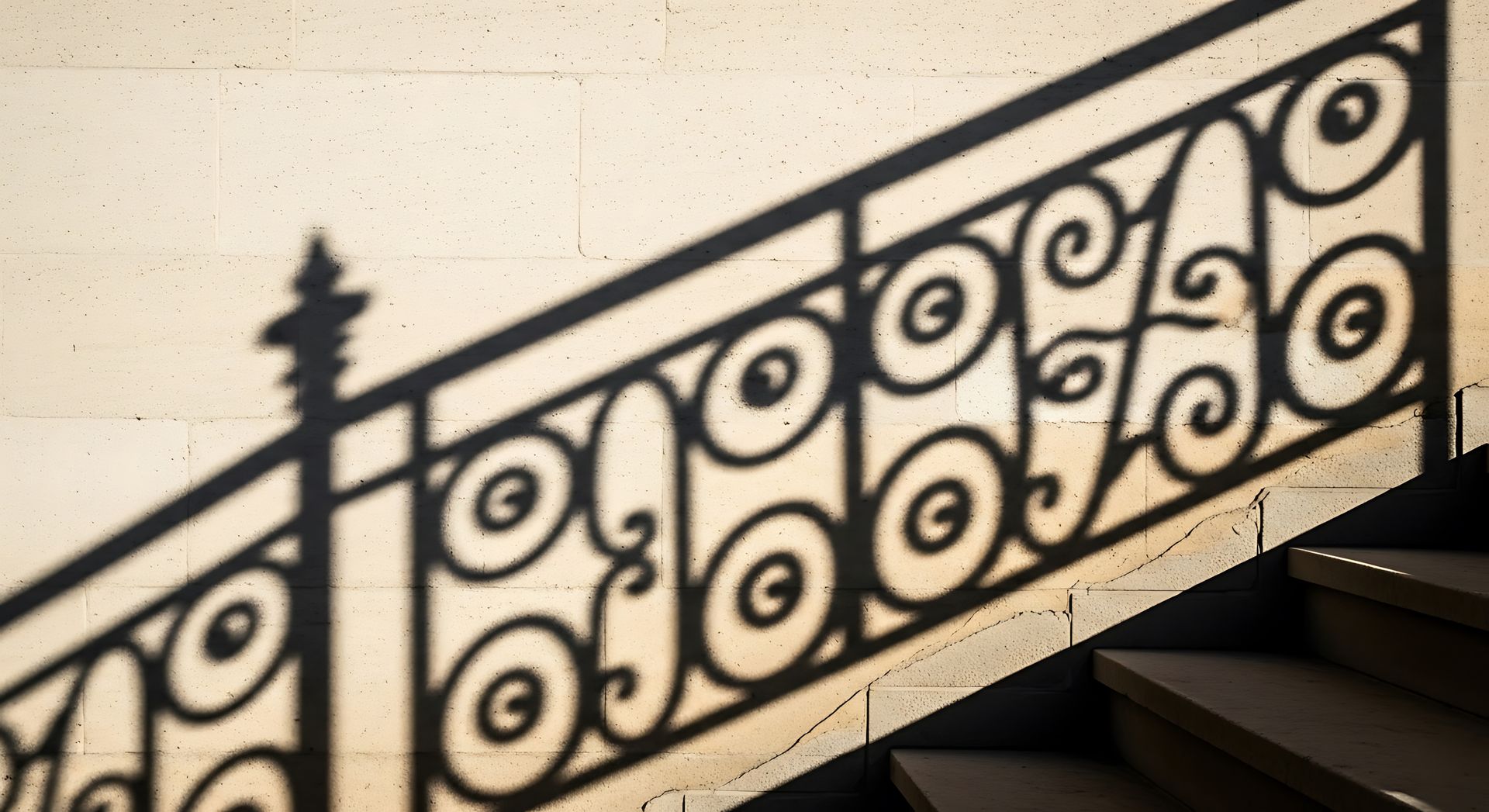 Une rampe d'escalier en fer forgé noir projette son ombre sur un mur beige au-dessus de marches sombres.