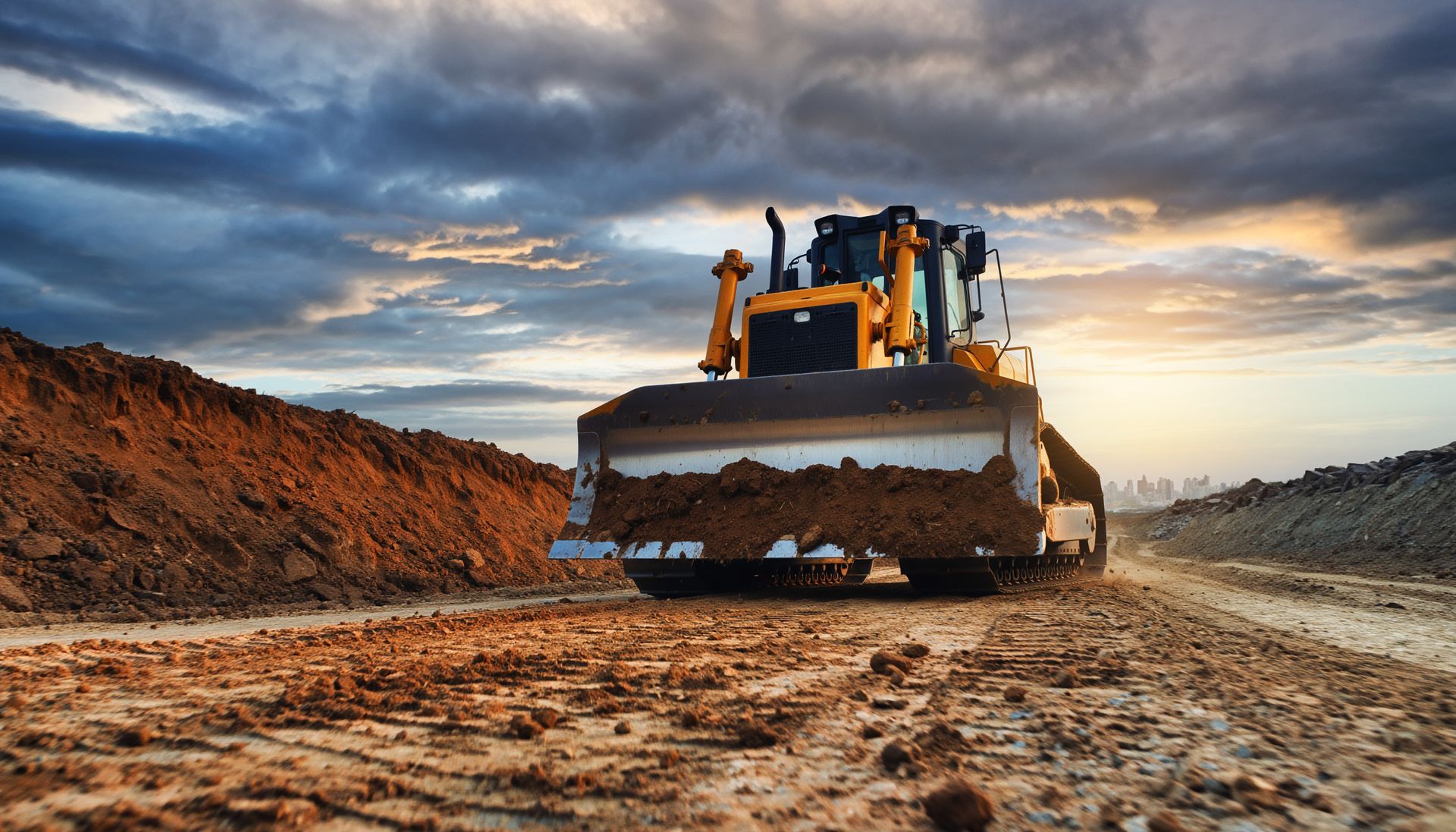 Un bulldozer nivelle un chemin de terre boueux au coucher du soleil sous un ciel nuageux.