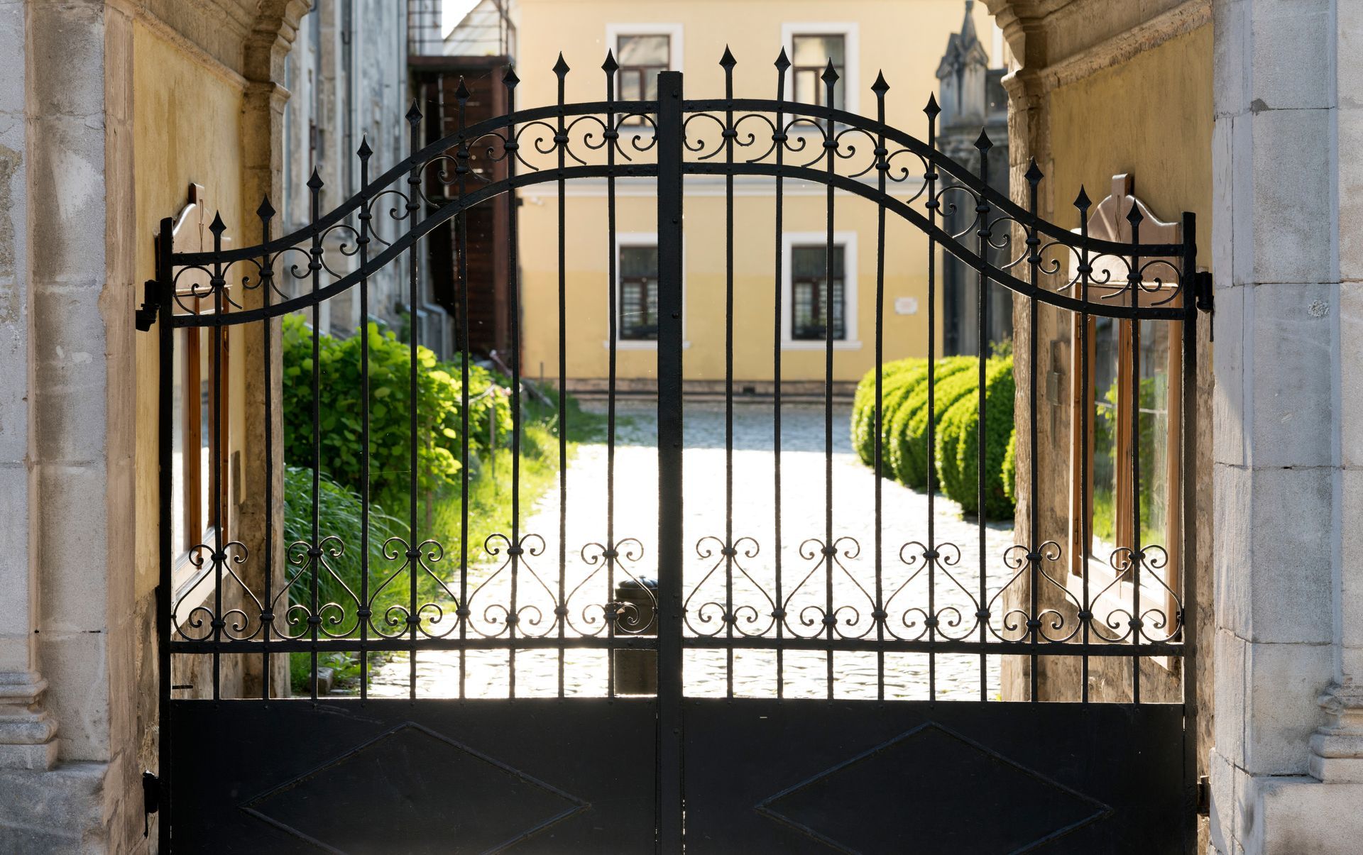 Un portail en fer forgé noir dans une cour en pierre, avec un chemin ensoleillé et un bâtiment beige au-delà.