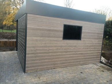Petit abri de jardin moderne marron avec un toit et une fenêtre noirs, sur une terrasse extérieure pavée.