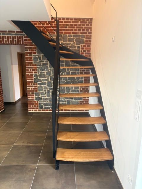 Escalier en bois courbé avec rampe noire, adossé à un mur de briques dans un intérieur moderne