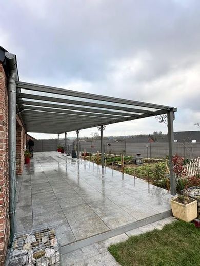 Patio couvert avec pergola métallique, sol carrelé humide et plantes en pot le long d'un mur de jardin