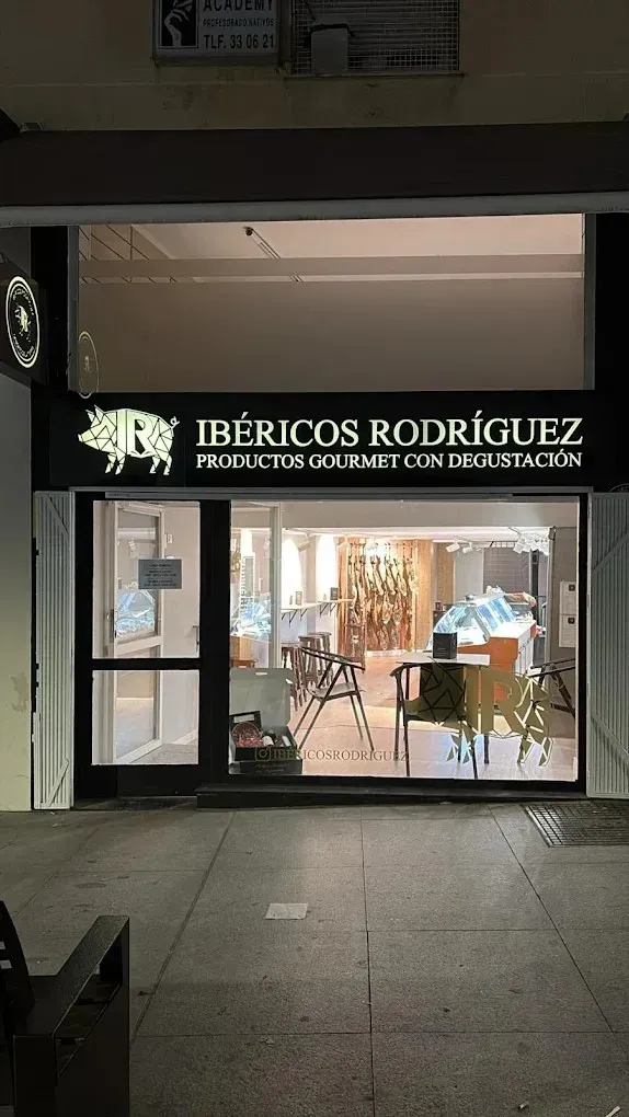 Fachada de la tienda de delicatessen Ibéricos Rodríguez con letrero iluminado y mesas visibles en el interior por la noche.