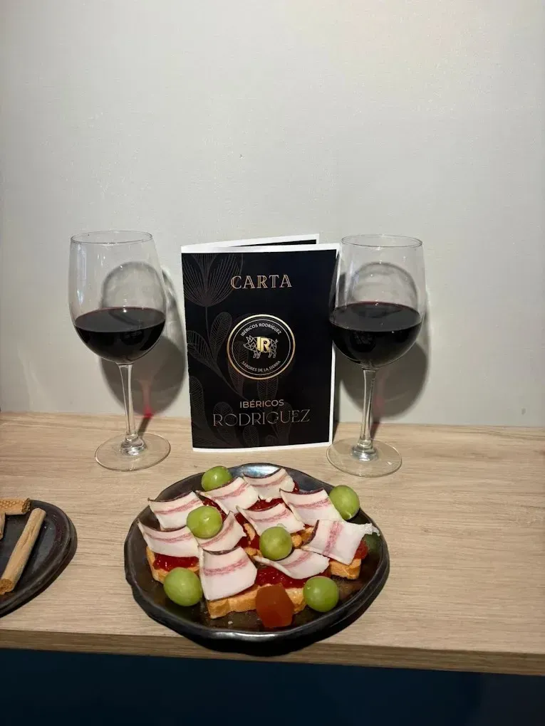 Copas de vino y una tabla de embutidos sobre una mesa con un menú al fondo.