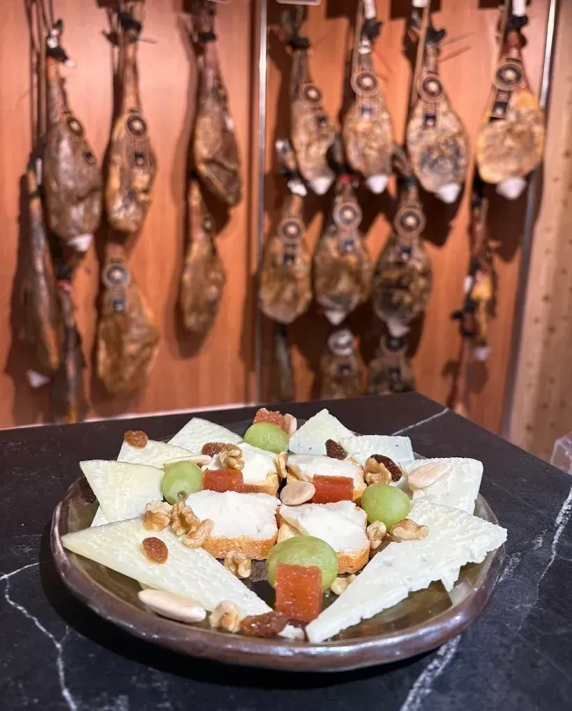Tabla de quesos con uvas y frutos secos en un plato, frente a embutidos colgados en un entorno rústico.