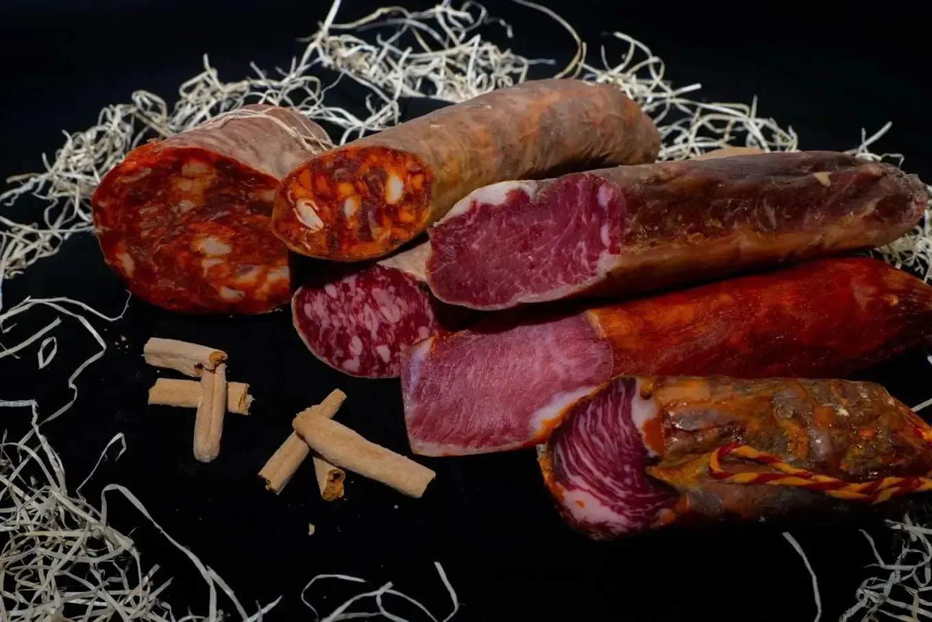 Salchichas curadas en rodajas con carne roja y marrón sobre paja y fondo oscuro.