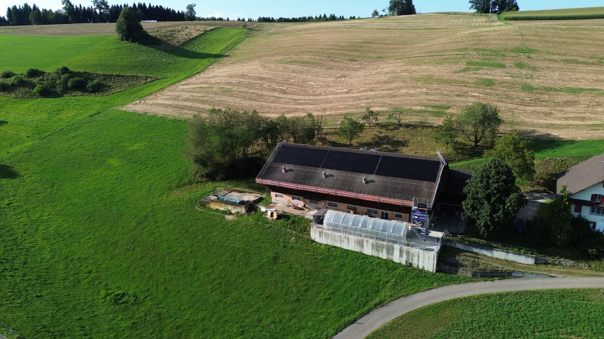Großes Bauernhaus mit Solardach inmitten grüner Hügel und Felder, umgeben von Bäumen und ländlicher 