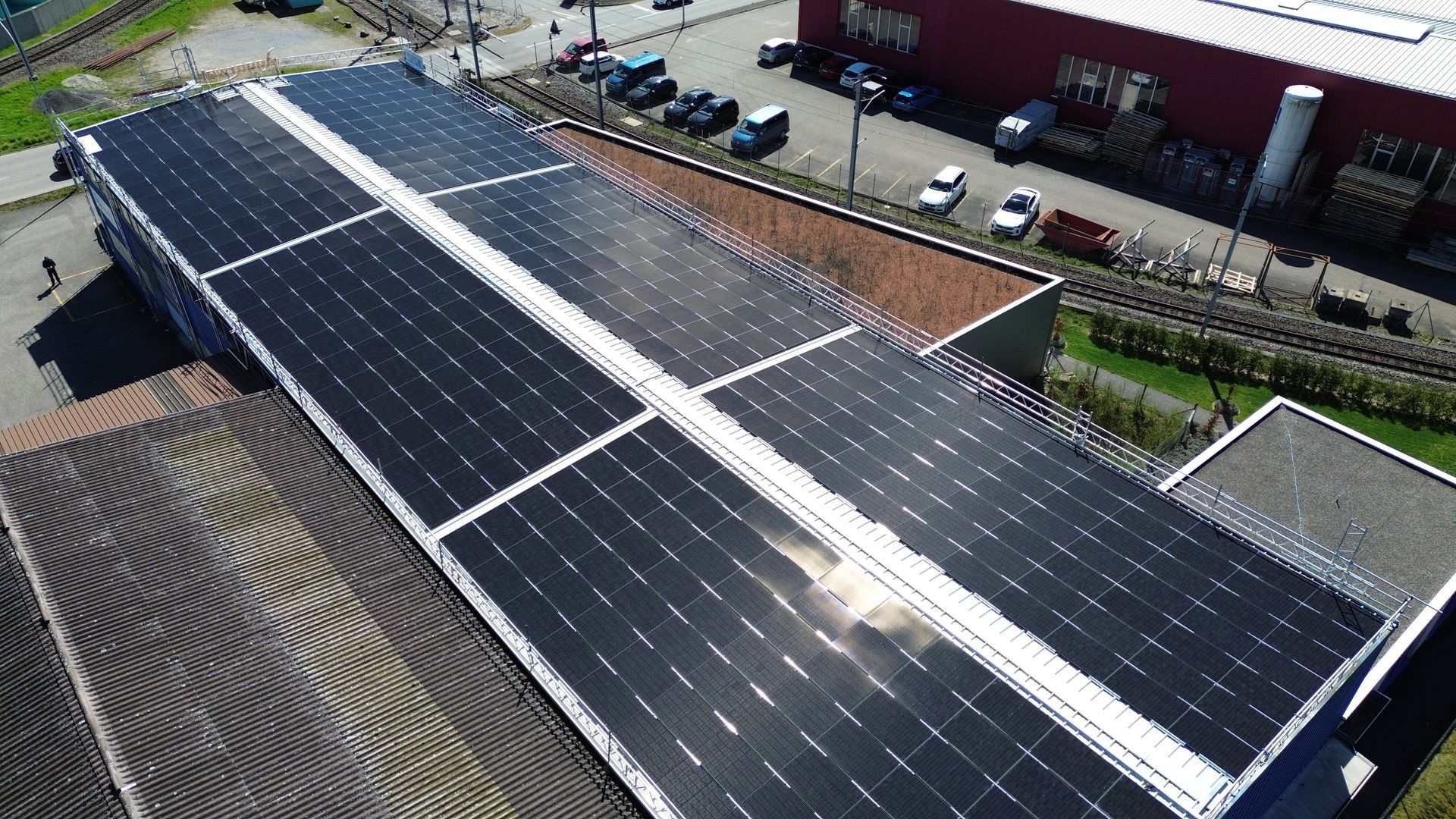 Industriehalle mit großer Solaranlage auf dem Dach, daneben Parkplätze mit Autos und Bahngeleise.
