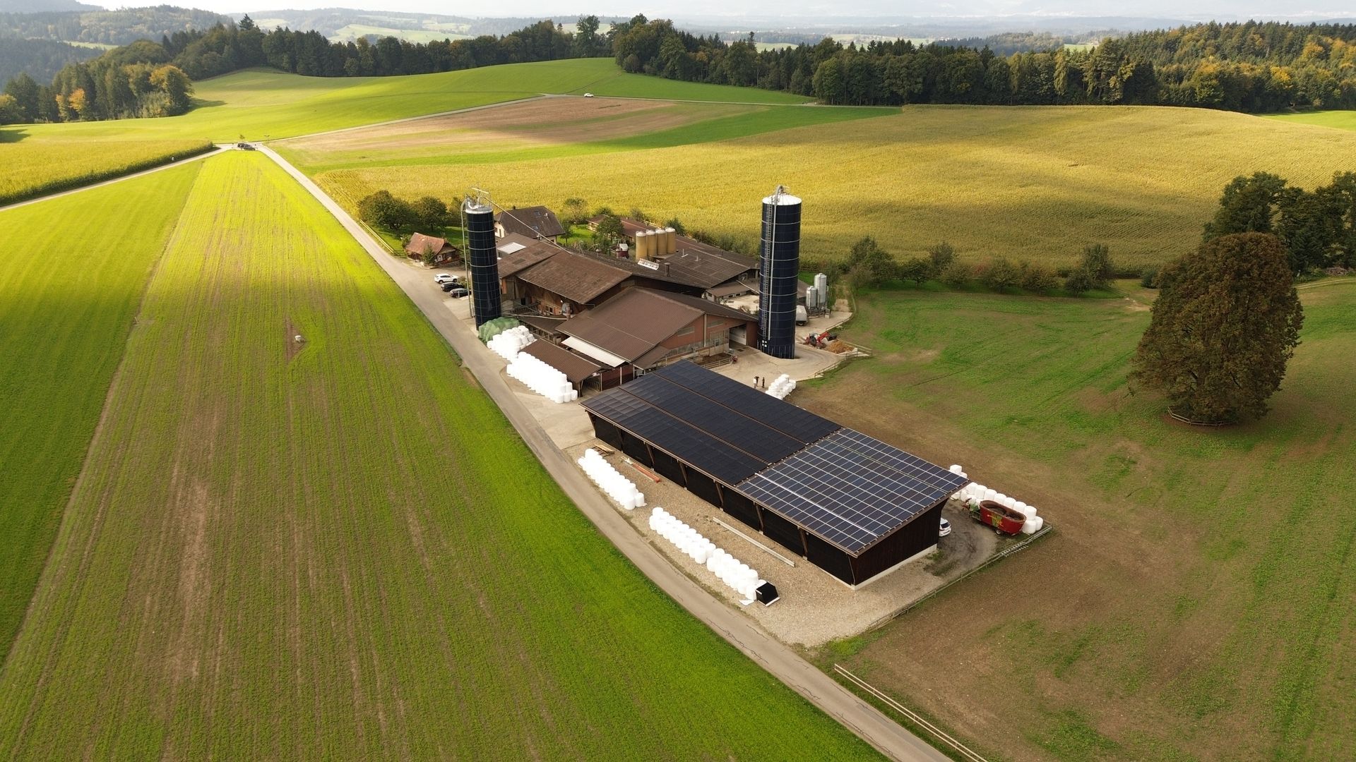 Luftaufnahme eines Bauernhofs mit Silos, Solarpanels auf dem Dach und grünen Feldern.