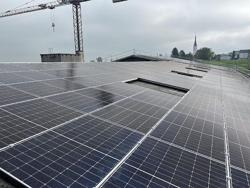 Solarpaneele auf einem Flachdach mit Baukran und Kirchturm im Hintergrund unter bewölktem Himmel