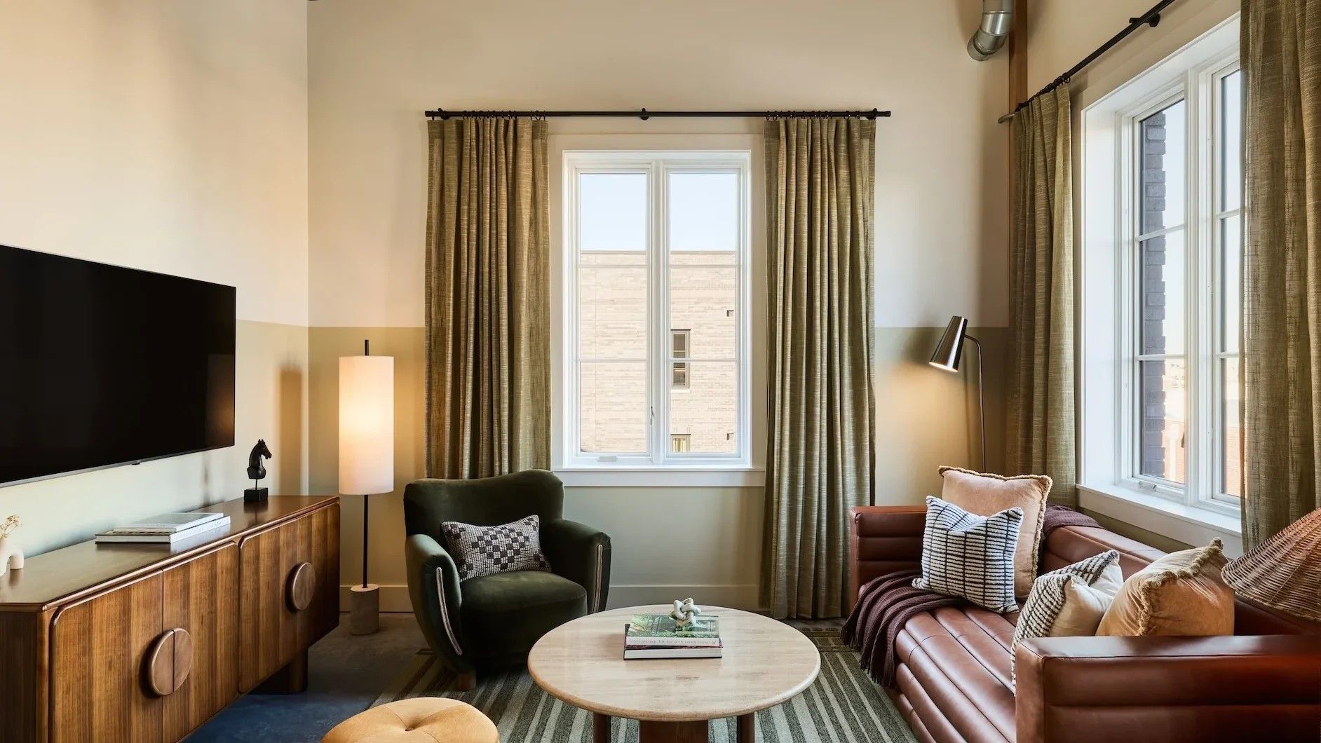 Living room with a sofa, armchair, and TV. Neutral walls, large windows with curtains.