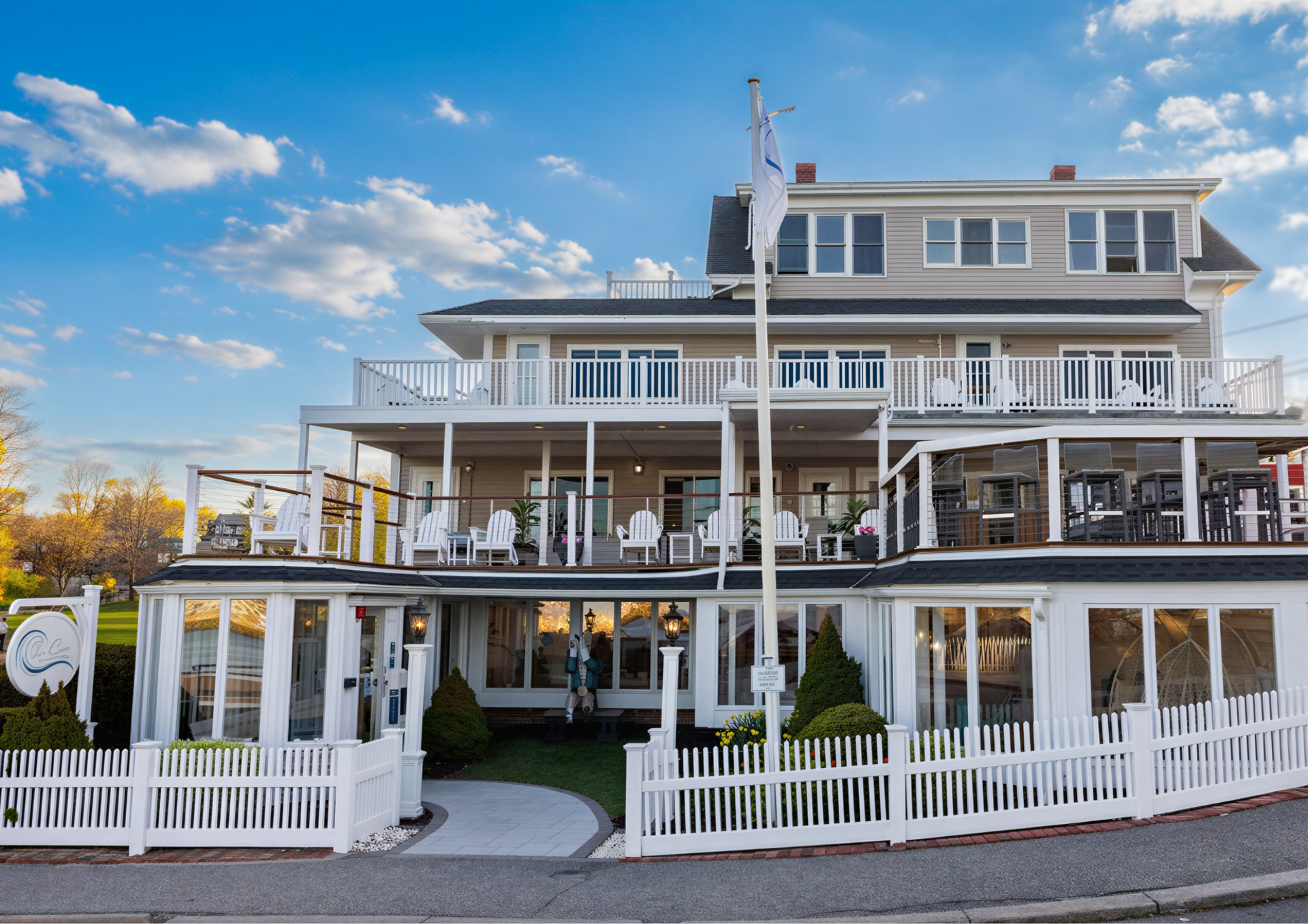 Large white waterfront building with wraparound porches and a white picket fence under a blue sky