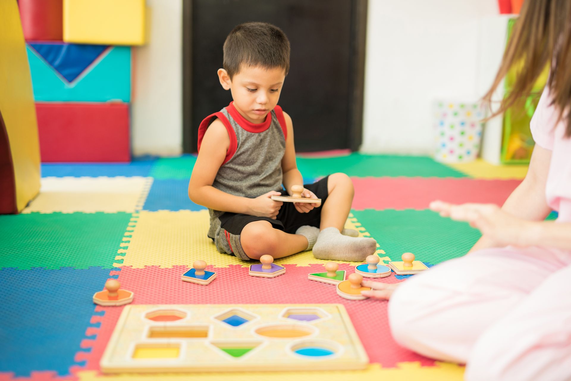 Un tout-petit joue avec un puzzle de tri de formes sur un tapis en mousse coloré tandis qu'un adulte est assis à proximité.