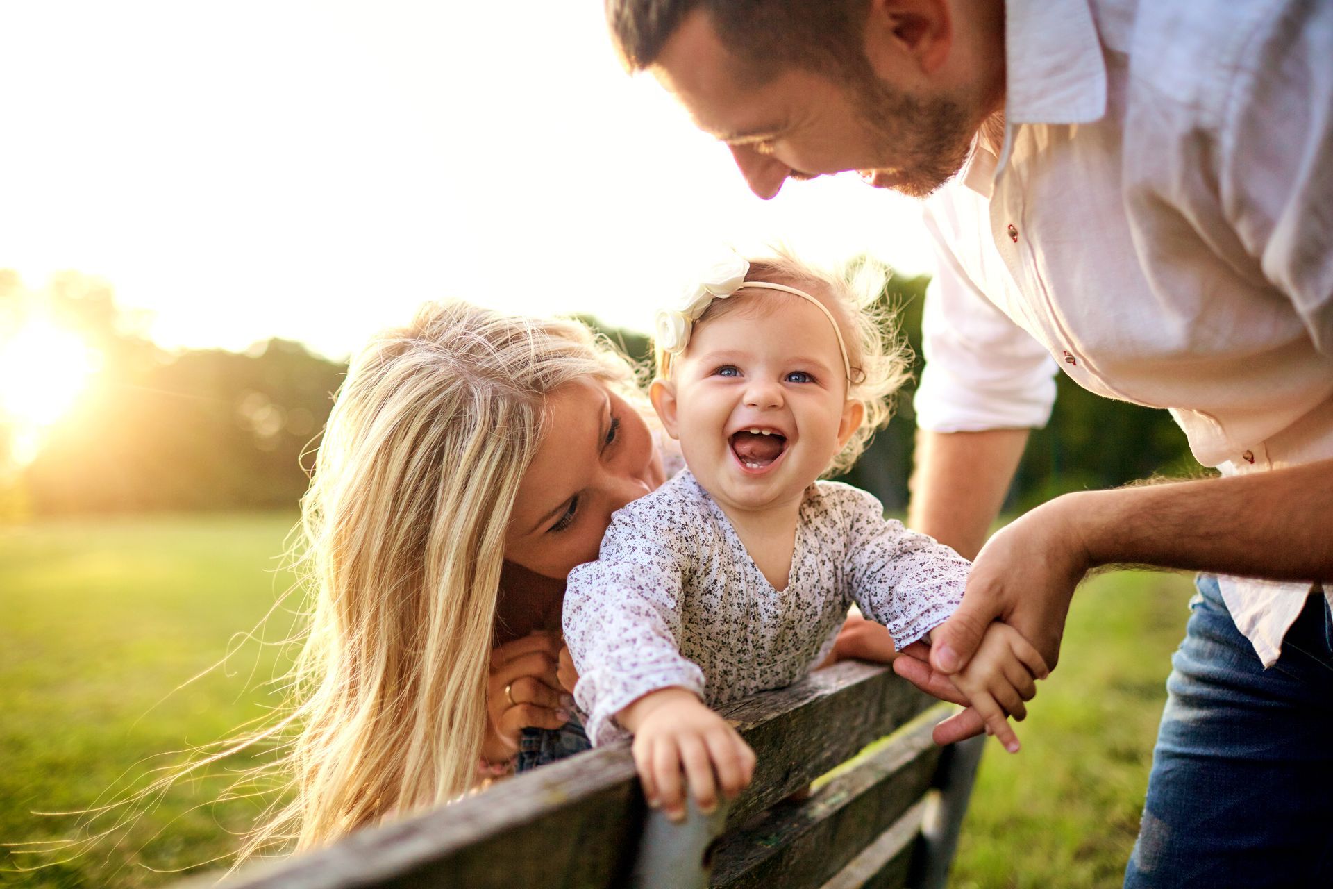 Une famille souriante, penchée par-dessus une clôture en bois dans un parc ensoleillé, avec un enfant riant au centre.
